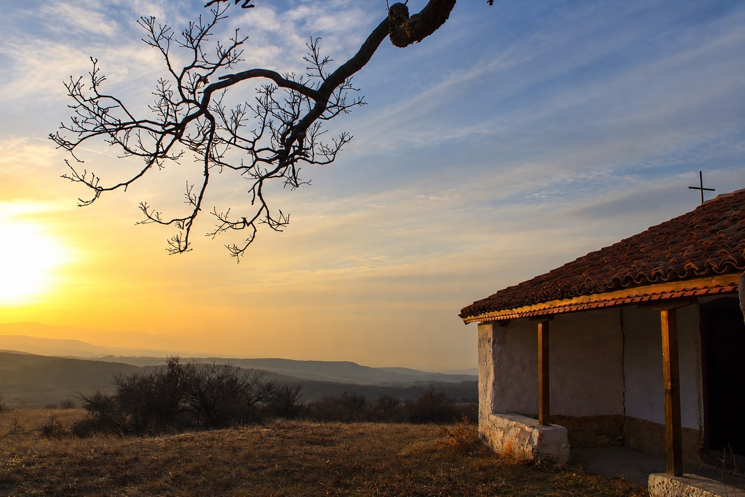 A small chapel on a hilltop at sunset, fields and distant hills below